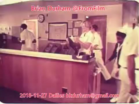 The still features a hospital or medical environment, showcasing staff in white uniforms. It depicts a reception area or nurse's station, with individuals engaged in tasks like walking or interacting with equipment. The overall atmosphere suggests a busy, organized setting typical of healthcare facilities. The date and credits indicate it is part of film dailies from November 27, 2018.