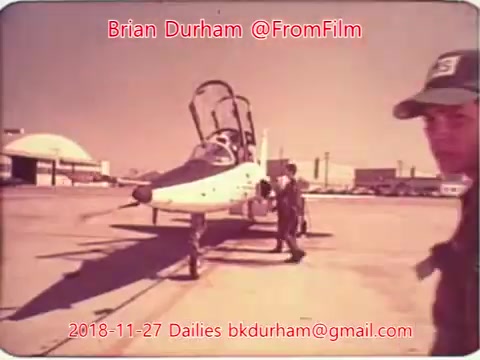 The still captures a scene on an airfield, featuring a person walking next to a small aircraft. The plane appears to have a distinctive cockpit design, and the background showcases airport hangars or buildings. The image is tinted with a vintage look, typical of 16mm film, suggesting it may be part of a historical or documentary-style collection. Text overlays provide information, including the date and contact details for the filmmaker.