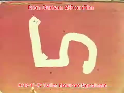 The still features a simple white painted design on a reddish background. The design resembles a loose, abstract letter 'L' or a squiggly line. It appears to be a frame from a film, possibly showcasing a stylized or artistic visual element. The image includes text credits related to Brian Durham and the date of the dailies.