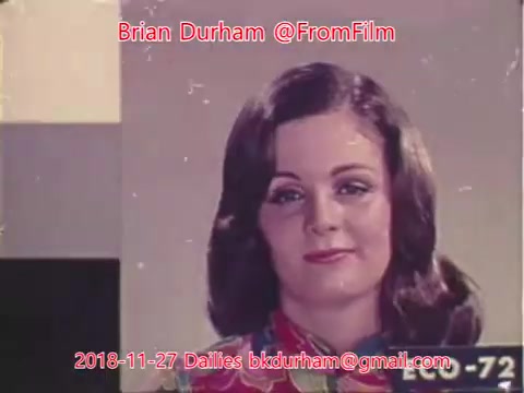 The still features a woman with long, flowing hair and a colorful patterned blouse. She appears to be in a studio setting, possibly for a recording or advertisement, and is looking directly at the camera with a confident expression. The image has a vintage quality typical of 16mm film, with some scratches or marks present. Text overlays include names and a date related to film dailies.