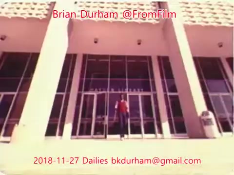 The still from '@FromFilm Dailies November 27, 2018' features a person standing in front of a modern, glass-fronted building with prominent pillars. The setting suggests a public space, likely an institutional or commercial establishment. The image has a vintage quality, typical of 16mm film, with a soft focus and warm tones. Text overlays provide context, including the creator's name and date.