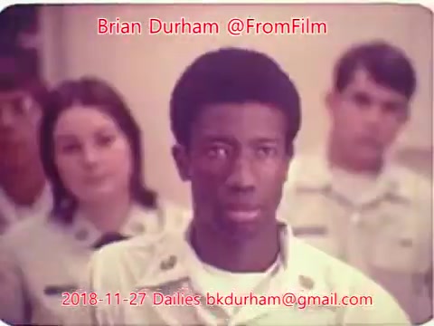 The still features a serious-looking young man in the foreground, with short black hair and wearing a light-colored shirt. Behind him are several other individuals, including a woman with long hair and a male figure in similar attire. The overall tone of the image suggests a moment of contemplation or focus. The context indicates it is from a film dailies collection dated November 27, 2018.