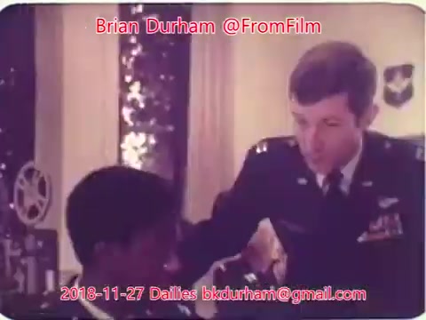 The still features two men engaged in a conversation. One man is wearing a military uniform, indicating a position of authority, while the other appears to be a civilian. The background includes elements suggesting an office or official setting. The image likely captures a moment from a film or television scene, reflecting themes of interaction and hierarchy.