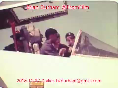 The still shows two people in the cockpit of an aircraft, wearing flight gear and engaged in conversation. One person is seated at the controls, while the other leans in, possibly giving instructions or discussing flight operations. The backdrop features the aircraft's dashboard and window. The image appears to capture a moment during a flight or pre-flight check. The date and contact information overlay adds context related to the footage source.