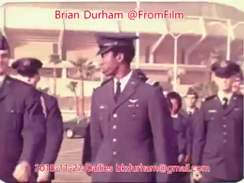 The still features a group of military personnel walking in uniform, likely at an outdoor location with a stadium in the background. The men are wearing dress uniforms and appear to be in formation, suggesting a ceremonial or official event. The colors are somewhat muted, typical of 16mm film, giving it a nostalgic quality.