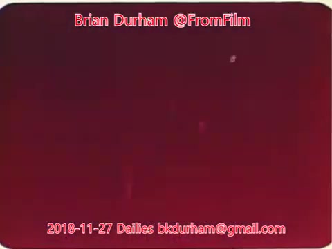 The still appears to be a deep red frame from a 16mm film, possibly showing a low-light or experimental scene. The composition includes some faint light spots, suggesting potential movement or depth. The text overlays indicate it is part of film dailies from November 27, 2018, related to a project by Brian Durham.