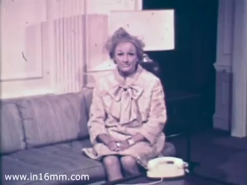 The still features comedian Phyllis Diller sitting on a couch in a well-furnished room. She wears a light-colored outfit with a large bow at the neckline, presenting a friendly demeanor. A retro-style telephone is visible on a small table beside her, and the decor suggests a mid-20th century aesthetic. The context is a 1968 public service announcement promoting the role of nurses in the Air National Guard.