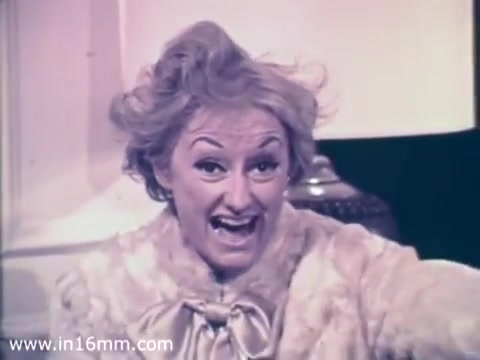 The still features Phyllis Diller, a well-known comedian, showcasing her signature animated expression and humorous demeanor. She's wearing a light-colored, fluffy garment, indicative of the period’s fashion, and is likely promoting the role of Air National Guard nurses. The background hints at a cozy, indoor setting, typical of 1960s public service announcements. The overall tone is upbeat and engaging, reflecting Diller's comedic style.