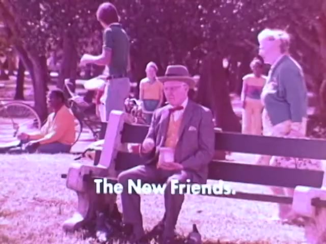 The still from the Kodak Instant Camera commercial features a park scene where various characters are engaged in activities. In the foreground, an elderly gentleman sits on a bench, appearing cheerful as he holds a cup. He is dressed in formal attire with a bow tie and a hat. Behind him, a group of people, including children and adults in colorful clothing, fills the background, suggesting a lively social atmosphere. The text 'The New Friends' is prominently displayed, highlighting the theme of connection through photography. The color palette leans towards pastel shades typical of the 1970s aesthetic.