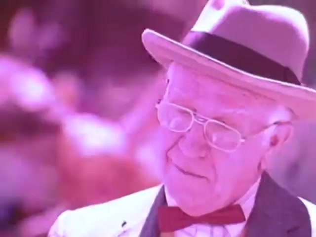 The still from the Kodak Instant Camera commercial features an elderly man dressed in a suit with a bowtie, wearing a light-colored hat. He appears to be engaged in a moment of reflection or observation, with a soft expression on his face. The color palette suggests a vintage feel, leaning towards pinkish tones, typical of 1970s film aesthetics. The overall scene likely conveys nostalgia and warmth, aligning with the themes of family and memorable moments captured by instant photography.