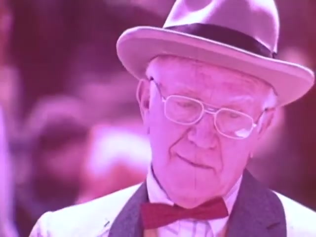 The still from the Kodak Instant Camera commercial features an older man in a light-colored suit and bow tie, wearing a fedora. He appears to be looking down thoughtfully, with a contemplative expression. The image has a pinkish hue, typical of the 1970s film aesthetic, creating a nostalgic atmosphere. This visual represents the approachable charm and emphasis on personal moments captured by the Kodak Instant Camera, highlighting its role in everyday life during that era.