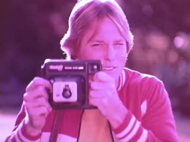 The still shows a young man wearing a red and white jacket, smiling as he holds up a Kodak Instant Camera. The image has a warm, nostalgic hue typical of 1970s aesthetics. The camera itself is compact and ergonomic, reflecting its design for ease of use. The outdoor setting hints at an informal, fun atmosphere, characteristic of family and social gatherings captured by instant photography.