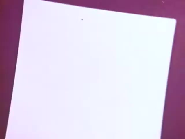 The still from the Kodak Instant Camera commercial features a close-up of a blank photo emerging from the camera's printing section. The image captures the anticipation of instant photography, highlighting the simplicity and immediacy of the Kodak Handle. The background is likely a vibrant color, emphasizing the excitement of capturing memories instantly. This commercial underscores the popularity of affordable instant cameras in the late 1970s and early 1980s.