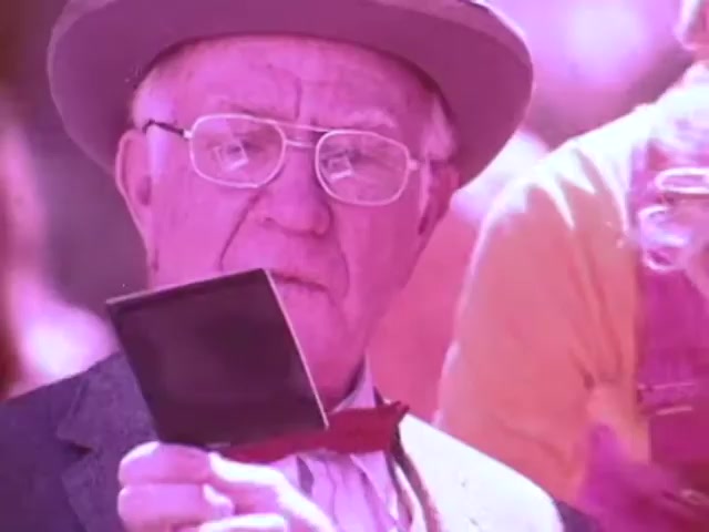The still from the Kodak Instant Camera commercial features an elderly man wearing a suit and a bow tie, closely inspecting a photo he just developed. He sports glasses and a hat, highlighting a classic, vintage look. The colors are notably vibrant, with a pinkish hue dominating the scene, giving it a nostalgic, retro appeal typical of the 1970s aesthetic. The focus is on the man's expression as he admires the instant photo, embodying the excitement and joy of capturing moments with the Kodak Handle.