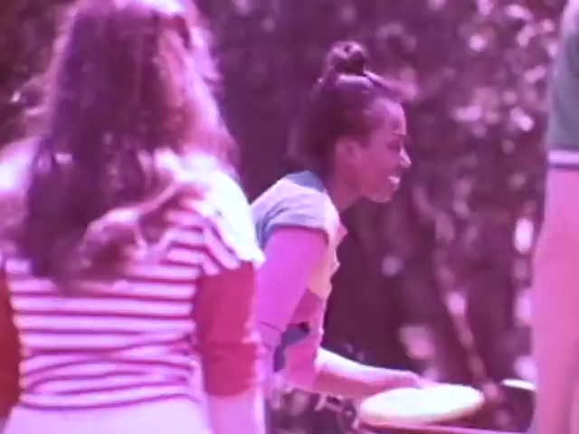 The still from the Kodak Instant Camera commercial features a vibrant outdoor scene from the late 1970s. A woman with a bun joyfully engages with a frisbee, emphasizing a fun, lively atmosphere. She wears a colorful top, while another woman in the background sports a striped shirt, suggesting a relaxed social gathering. The overall color tone has a warm, slightly pinkish tint, typical of retro film aesthetics, enhancing the nostalgic feel of the era.