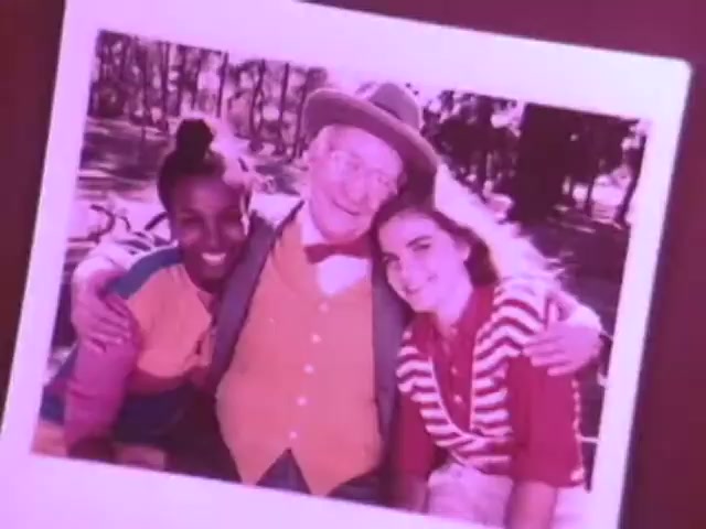 The still from the Kodak Instant Camera commercial features a cheerful group of three individuals posing closely together, smiling broadly. The image appears to be printed in vibrant colors typical of instant photography from the 1970s. The setting seems to be outdoors, likely in a park, with dappled sunlight and trees in the background. This commercial highlights the accessibility and fun of capturing spontaneous moments with the Kodak Handle instant camera.