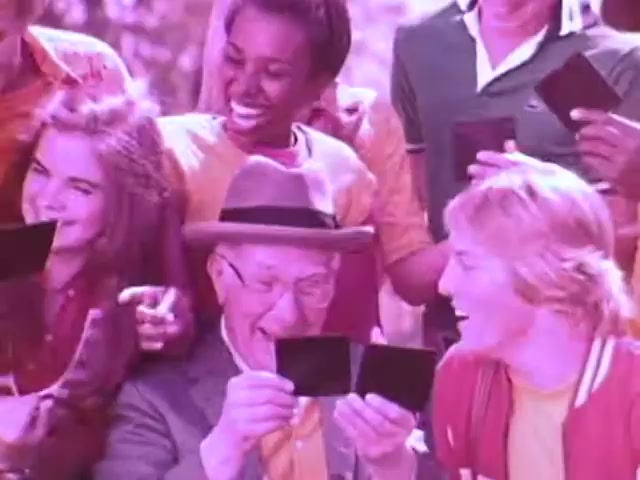 The still from the Kodak Instant Camera commercial features a diverse group of people joyfully engaging with their instant photos. Centered is an elderly man wearing a hat, smiling broadly as he holds up a photograph, surrounded by excited young individuals. They display their own photos, reflecting a shared moment of fun and nostalgia. The overall aesthetic has a warm, nostalgic tint, characteristic of the 1970s, emphasizing the camera's accessibility and appeal.