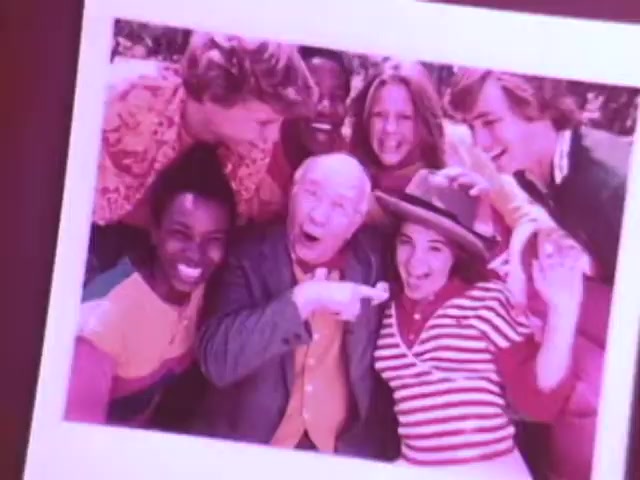 The still from the Kodak Instant Camera commercial features a joyful group of six people gathered together, posing for a photograph. They display a range of expressions, from smiles to playful gestures, creating a lively atmosphere. The scene captures the essence of nostalgia with its vibrant colors and fashion typical of the 1970s. The image promotes the fun and social aspects of using the Kodak Handle instant camera, emphasizing its ability to capture memorable moments instantly.