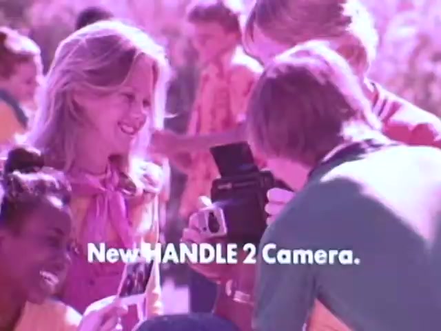 The still from the Kodak Instant Camera commercial features a group of cheerful young people outdoors, showcasing the Kodak Handle 2 camera. A boy is demonstrating the camera while a girl smiles, holding a freshly developed photo. The vibrant colors and playful atmosphere of the 1970s suggest a carefree, fun moment, emphasizing the joy of capturing memories with this accessible instant camera. The text highlights the 'New HANDLE 2 Camera' prominently, appealing to a youthful audience.