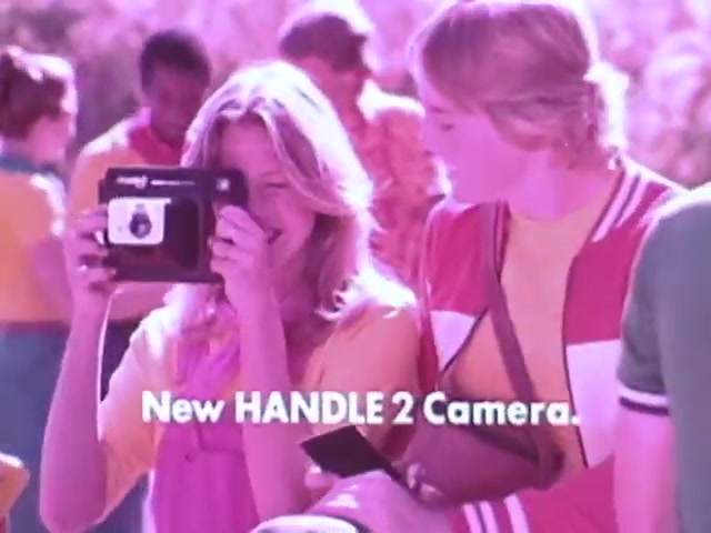 The still from the Kodak Instant Camera commercial features a cheerful young woman smiling as she holds the Handle 2 camera to take a photo. She's dressed in a colorful outfit, reflecting the vibrant 1970s style. Next to her, a friend looks on, also in bright attire. The background shows a playful group of people, reinforcing the fun atmosphere. The text at the bottom announces 'New HANDLE 2 Camera,' highlighting the product's appeal as a modern, accessible instant photography option.