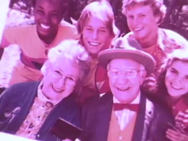 The still from the Kodak Instant Camera commercial features a cheerful group of five individuals posing closely together. In the foreground, an elderly couple smiles widely, with the woman holding a Kodak camera. Behind them, three children, all smiling, represent a sense of joy and togetherness. The vibrant colors and casual attire reflect the playful spirit of the 1970s, highlighting the appeal of Kodak's affordable instant camera. The scene captures a moment of familial bonding and the fun of instant photography.