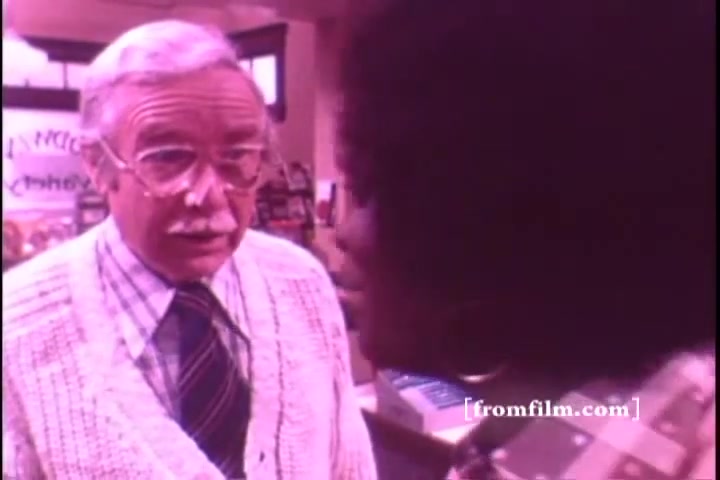 The still shows two characters engaged in conversation in a setting likely resembling a store or pharmacy. One character, an elderly man with glasses and a sweater vest, appears to be listening intently, while the other, a young person with an afro hairstyle, is animatedly speaking. The scene conveys a friendly yet serious interaction, possibly discussing the benefits of Crest toothpaste. The overall color tint suggests an older film quality typical of 16mm prints.