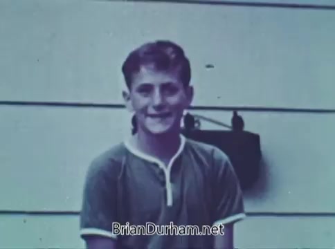 The still features a young boy smiling at the camera, dressed in a simple, short-sleeved shirt with a white collar detail. He stands against a backdrop of a wooden structure, likely a house, which has a plain exterior. The image is likely from a 1968 public service announcement by Save the Children, emphasizing children’s issues and welfare. The boy’s expression conveys innocence and positivity, aligning with the PSA's mission.