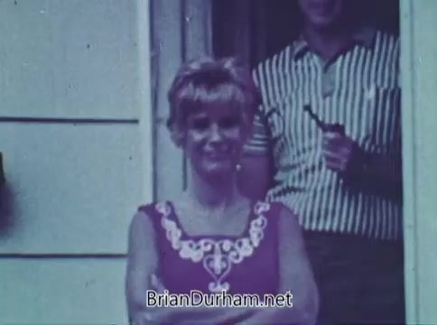 The still captures a woman with blonde hair, smiling confidently while standing in a doorway. She wears a sleeveless, vibrant purple dress adorned with white embroidery. In the background, a man partially visible seems to be holding an object, likely a beverage. The image evokes a casual, domestic setting typical of the late 1960s, contributing to the overall theme of a public service announcement.