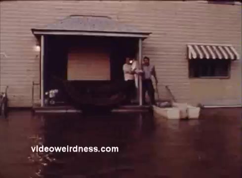 The still depicts a scene where two individuals stand on a porch, surrounded by floodwaters. The building features a light-colored exterior with a striped awning over one of the windows. There are containers on the ground, likely for storing items away from the water. The atmosphere suggests a serious situation, likely aimed at raising awareness about flooding and its impact on the community, fitting within the context of a public service announcement by Montana Fish and Game.