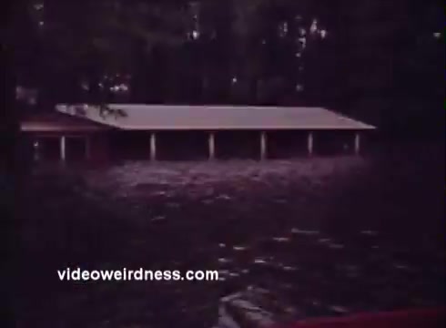 The still from the 'Montana Fish and Game PSA' depicts a flooded area, with a structure partially submerged in water. The setting appears rural and wooded, contributing to a sense of environmental impact. The image likely serves to raise awareness about the consequences of flooding on local wildlife and habitat.