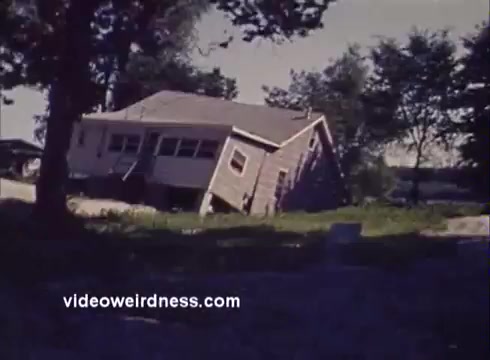 The still from the 'Montana Fish and Game PSA' depicts a house that appears to be leaning or partially collapsed, surrounded by greenery. The scene suggests a sense of dislocation or distress in a natural setting, possibly emphasizing issues related to environmental impact or safety in the region. The visual composition captures a moment of unsettling contrast between the structure and its surroundings.