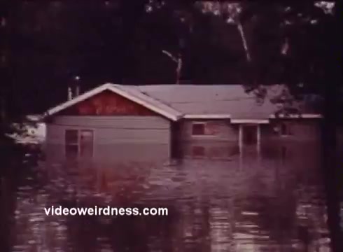 The still from the 'Montana Fish and Game PSA' depicts a house partially submerged in floodwater. The structure appears to be in a rural setting, surrounded by trees. The water level rises significantly, emphasizing the impact of flooding. The image serves as a visual warning, likely related to the effects of natural disasters on wildlife and habitat in Montana.