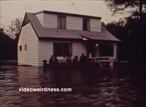 The still depicts a house partially submerged in water, surrounded by flooding. A group of people can be seen on the porch, likely seeking refuge. The scene conveys a sense of urgency and concern for safety, aligning with the PSA's message about wildlife and environmental issues in Montana. The muted colors reflect the somber mood of the situation.
