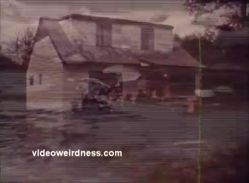 The still depicts a small, distressed house partially submerged in floodwaters, capturing a moment likely related to environmental awareness. The scene conveys a sense of urgency and highlights the consequences of natural disasters, aligning with the themes of conservation and public safety in the Montana Fish and Game PSA. The visual quality suggests a vintage or retro film aesthetic.