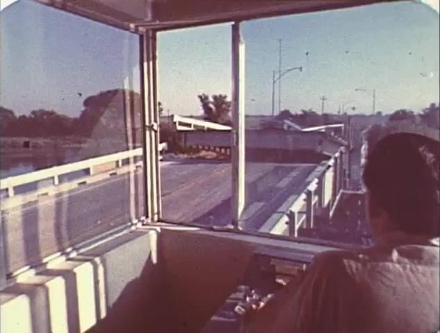 The still features a perspective from inside a vehicle, showing a view through a large window. The landscape includes an open road, with road signs visible in the distance. A person can be seen from behind, seemingly focused on driving. The lighting suggests it may be a sunny day, highlighting the scene's sense of movement. This image likely connects to the themes of speed and adventure prevalent in the films 'Dirty Mary, Crazy Larry' and 'Vanishing Point.' Given the context of a TV spot trailer, it aims to evoke excitement around the action and high-stakes driving featured in both films.
