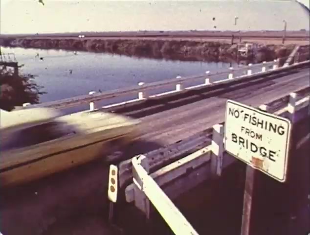 The 16mm still from the TV spot trailer for 'Double Bill: 'Dirty Mary, Crazy Larry' and 'Vanishing Point'' depicts a scene featuring a fast-moving yellow car on a bridge. A sign reading 'NO FISHING FROM BRIDGE' is prominently displayed on the right. The location appears to be rural, with a body of water visible next to the bridge, indicating a serene yet dynamic environment. This imagery encapsulates the high-speed themes characteristic of both films, highlighting the thrill of car chases and the sense of freedom associated with open roads. The overall aesthetic suggests a vintage look, typical of trailers from that era.