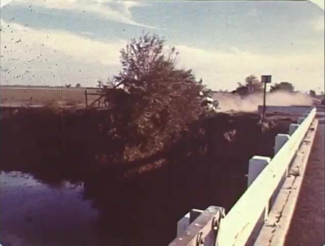 The 16mm still from the TV spot trailer for 'Double Bill: 'Dirty Mary, Crazy Larry' and 'Vanishing Point'' depicts a dusty scene featuring a tree beside a roadway. The setting suggests a rural landscape, likely hinting at the high-speed car chases that are central to both films. The inclusion of a guardrail and a sign indicates a tension or impending action, a common element in trailers aimed at highlighting the thrill and excitement of the featured films. This reissue trailer combines classic elements from both movies to attract fans of the genre.