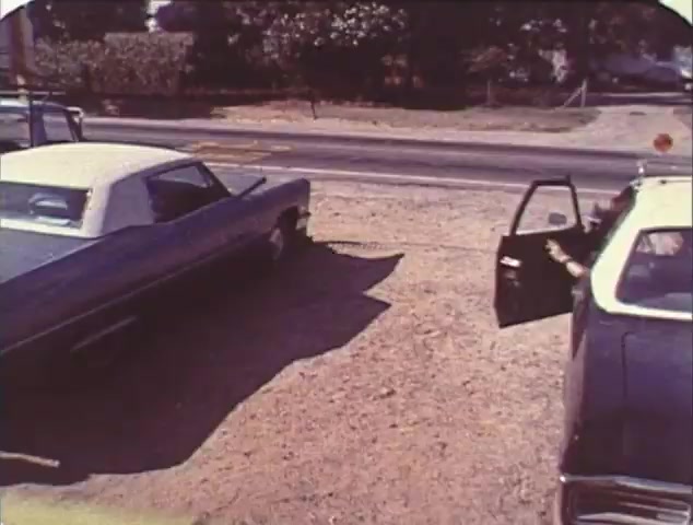 The still from the 'Double Bill: 'Dirty Mary, Crazy Larry' and 'Vanishing Point'' TV spot trailer features a parking lot scene with two classic cars, one in blue and the other in black, parked beside a road. The driver’s side door of the black car is open, suggesting a moment of action or transition. The setting appears sunny, hinting at a vintage aesthetic aligned with the films' themes of car culture and escapism. This trailer likely serves as a nostalgic reissue, promoting the pairing of both films known for their high-octane chases.