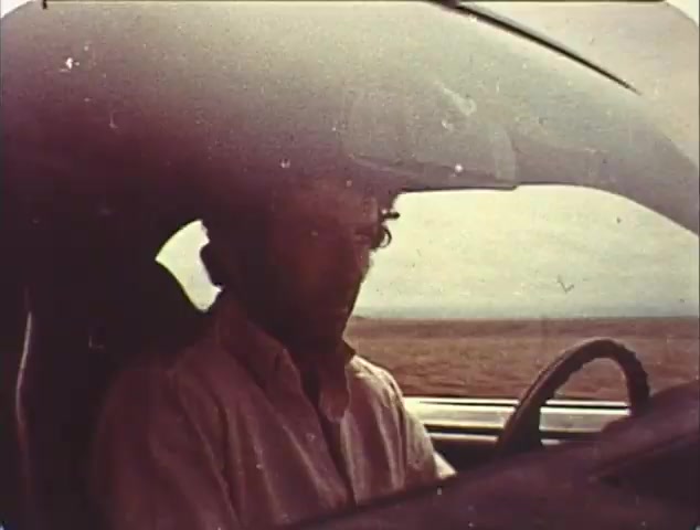 The still from the 16mm trailer for 'Double Bill: 'Dirty Mary, Crazy Larry' and 'Vanishing Point'' focuses on a character inside a car, seated in the driver's seat. The environment outside the window suggests a vast, open landscape. The image has a vintage feel, with a slightly faded quality typical of older film stock, capturing a moment likely filled with tension or anticipation. This reissue trailer aims to evoke nostalgia for the classic films it promotes.