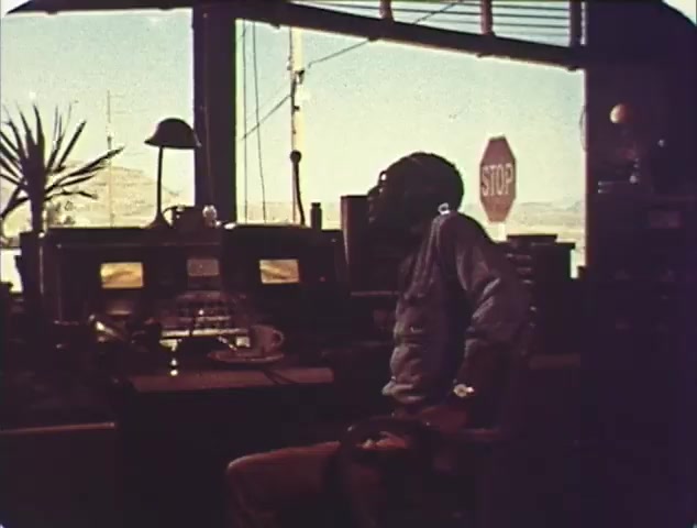 The still from the 'Double Bill: Dirty Mary, Crazy Larry and Vanishing Point' TV spot trailer shows a lone figure seated at a control station, surrounded by a cluttered room filled with equipment. Large windows reveal a bright, expansive landscape outside, with a visible stop sign in the background, hinting at themes of road and control. The overall tone is contemplative, possibly foreshadowing the action-packed elements of the films featured in the trailer. The color palette suggests a vintage feel, typical of 16mm film aesthetics.