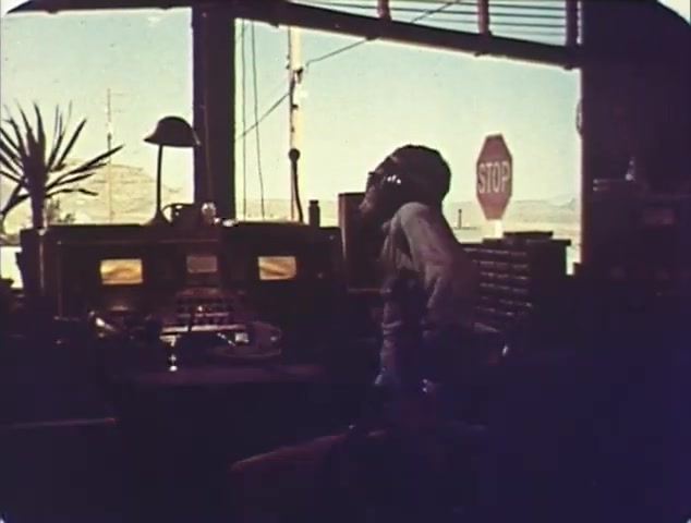 The still from the reissue trailer for 'Double Bill: 'Dirty Mary, Crazy Larry' and 'Vanishing Point'' features a scene set in a control room or office, likely connected to highway patrol or transportation. The focus is on a figure in a shirt, seemingly engaged in communication, with a stop sign visible through a window, hinting at themes of traffic and urgency. The overall lighting conveys a vintage atmosphere, consistent with 16mm film quality, enhancing the retro aesthetic of the trailers.