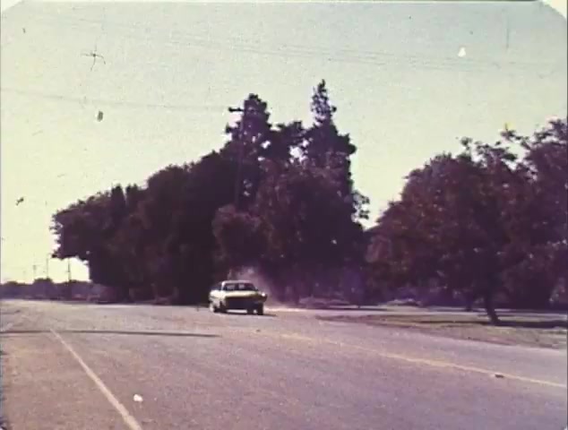 The still shows a classic car driving down a rural road, surrounded by dense greenery and trees. The scene appears to be set in broad daylight, emphasizing an open and expansive landscape. This still likely captures the essence of high-speed pursuits common in the films 'Dirty Mary, Crazy Larry' and 'Vanishing Point,' reflecting the themes of freedom and adventure prevalent in both movies, which are featured in the Double Bill trailer. The vintage 16mm quality adds a nostalgic touch to the promotional material.