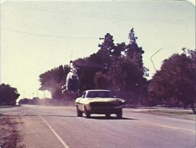 The still from the TV spot trailer for 'Double Bill: 'Dirty Mary, Crazy Larry' and 'Vanishing Point'' depicts a dramatic action scene featuring a yellow car speeding down a road as a helicopter hovers above. The road is flanked by trees, suggesting an open, rural setting. The image captures the high-stakes excitement typical of the films, showcasing thrilling car chases and aerial maneuvers. The 16mm quality gives it a vintage look, characteristic of the era's film production.