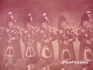 The still features a group of men dressed in traditional Scottish attire, including kilts and feathered hats, playing bagpipes. They are arranged in a line against a misty, ethereal background, creating a celebratory and nostalgic atmosphere, reminiscent of 1970s advertisements for Seagram's 100 Pipers Scotch. The color tones appear soft and slightly faded, characteristic of a 16mm print.