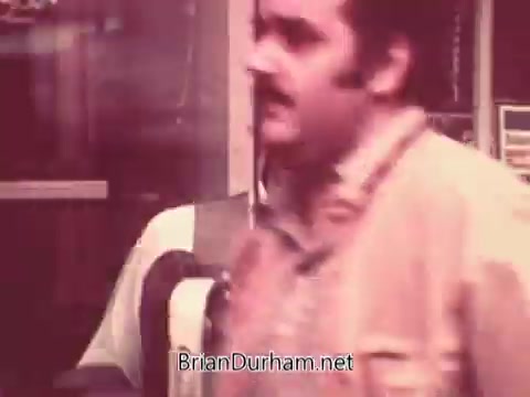 The still from 'Ohio Job Service PSA 3 - 1970s' features a man with a mustache, dressed in a light-colored shirt. He appears to be in an urban setting, possibly outside a job service office, with signs and a storefront visible in the background. The image has a vintage sepia tone, typical of 16mm film from that era, suggesting a focus on job-seeking and employment services. The context implies a public service announcement aimed at aiding job seekers in Ohio during the 1970s.