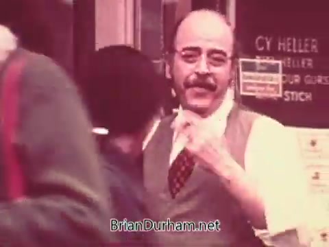 The still shows a man with glasses and a mustache, dressed in a vest and tie, interacting with passersby in a busy street scene. Various people are visible in the background, with signs indicating a storefront. The atmosphere reflects a lively 1970s urban environment, likely promoting job services.