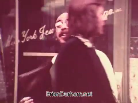 The still depicts two people interacting outside a storefront, likely part of a public service announcement. One person appears to be engaged in conversation with a cheerful demeanor, while the other, with longer hair, seems to be walking away or turning. The background features a window display that includes text, possibly indicating the location or theme of the PSA, which is related to job services in Ohio during the 1970s. The overall tone seems informal and engaging.