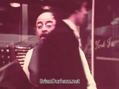 The still from 'Ohio Job Service PSA 3 - 1970s' features a busy scene likely set in an office or job placement environment. A man in a suit is prominently seen in the foreground, animatedly engaging with another individual who is partially cropped out of the frame. This snapshot captures a moment of interaction, hinting at themes of employment and job searching relevant to the period. The overall color tone is warm, typical of archival footage from that era.