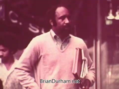 The still features a man walking along a city street, holding several books under his arm. He is wearing a light sweater over a collared shirt, giving a casual yet professional appearance. In the background, another individual can be seen, adding to the urban setting. The overall color tone of the image has a warm, somewhat faded quality typical of 1970s film.