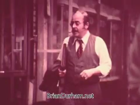 The still from 'Ohio Job Service PSA 3 - 1970's' features a man in a suit and tie, walking outside a building. He appears to be middle-aged, with a mustache and a briefcase in hand. The image has a slightly faded, vintage quality typical of 16mm film, reflecting the era of the 1970s. The background includes a windowed structure, suggesting an urban setting. The overall tone conveys a professional atmosphere related to job services.